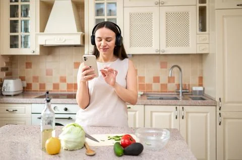 Woman using smartphone while preparing vegetables in kitchen. Wearing headpho Stock Photos