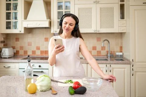 Woman using smartphone while preparing vegetables in kitchen. Wearing headpho Stock Photos