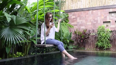 Woman using smartphone while sitting on swing near pool in tropical country. Stock Footage 75031091