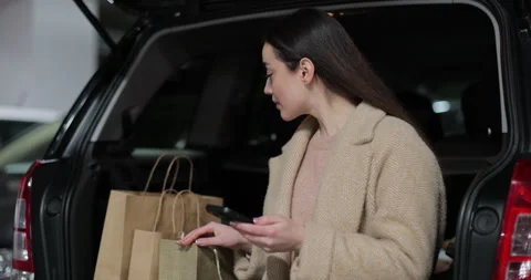 Woman using a smartphone while sitting in the trunk of a car in shopping center Stock Footage 297755274