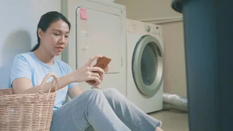 Woman Using Smartphone While Sitting Next to Washing Machine Stock Footage 314786789