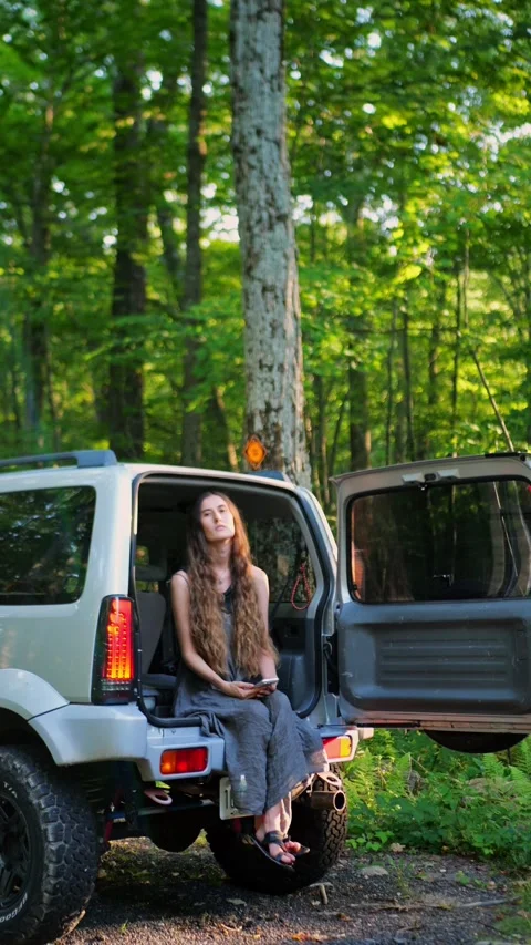 Woman using smartphone while sitting in open SUV trunk in sunlit forest... Video stock 340291022