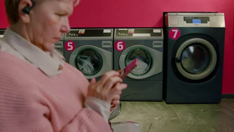 Woman using smartphone while waiting in selfservice laundry facility with modern Stock Footage 329366253