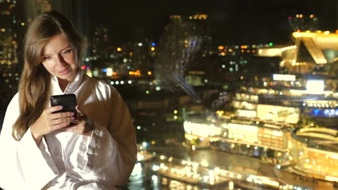 Woman using smartphone by window with night city view. Urban lifestyle and Видео 260336908