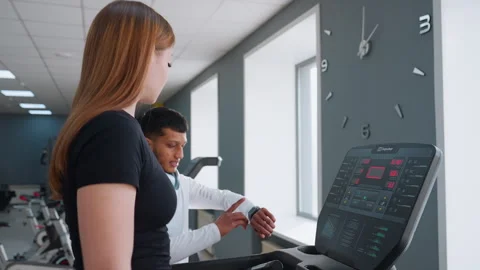 Woman Using Treadmill While Trainer Monitors Time in Modern Fitness Gym Stock Footage 308320004
