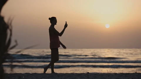 Woman using video chat while walking on the beach at sunset 스톡 동영상 104920096
