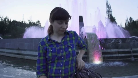 A woman is using a white smartphone next to a fountain in the evening Stock Footage 79137227
