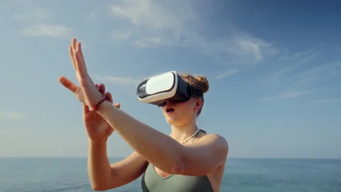 Woman in VR headset explores virtual beach scene, gesturing with hands. Female Stock Footage 264070308