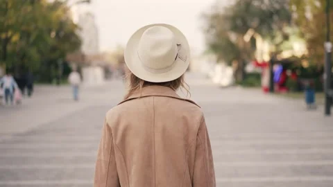 Woman walking down a tree-lined path in a beige coat and hat on a sunny day Stock Footage 293945454