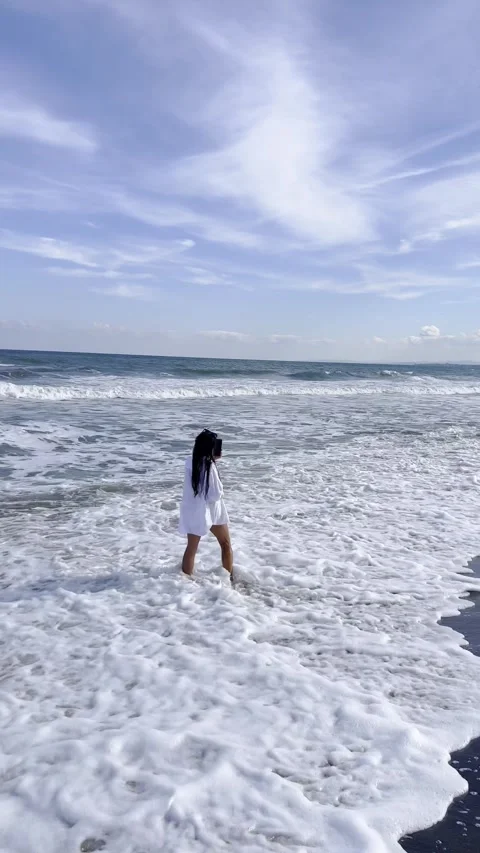 Woman walking into sea foam while filming ocean on smartphone Video stock 344536997