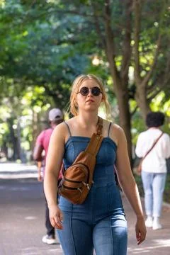 Woman walking on tree-lined path, wearing denim jumpsuit, round sunglasses, Stock Photos