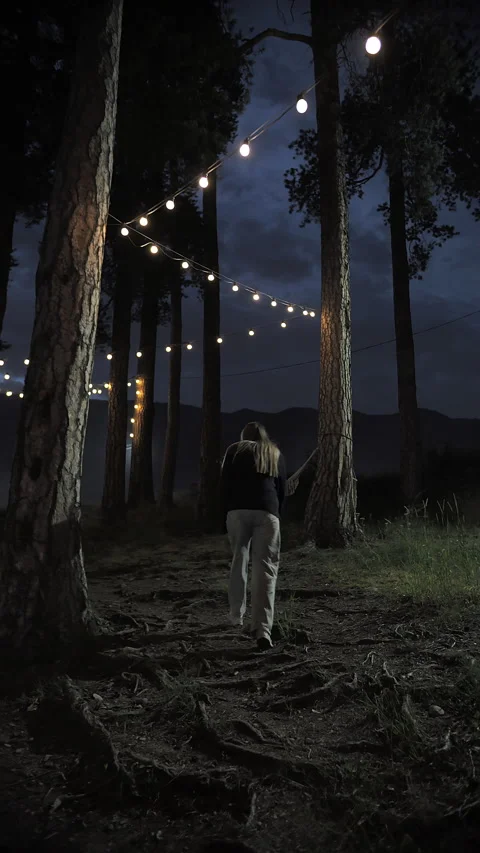 Woman walking under string lights in a forest at night Stockbeeldmateriaal 284336393