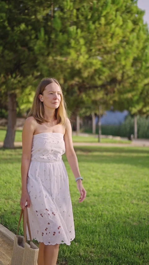Woman walks in park wearing light dress and holding bag on sunny day Stock Footage 346016853