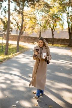 Woman walks on a path while using a phone and holding a drink in autumn setti 스톡 사진