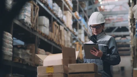 Woman in warehouse in helmet and gray uniform opens a cardboard box Stock Footage