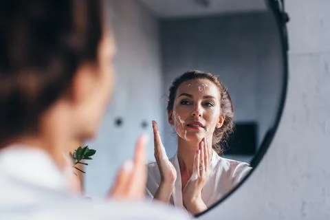 Woman washes in front of the mirror, applying foam to her face Stock Photos
