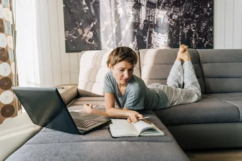 A woman watches notes in a notebook, using a laptop lying on the couch 스톡 사진