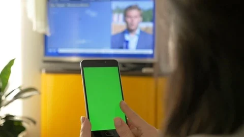 A woman is watching TV, and is holding a smartphone with a green screen. On the Stock Footage