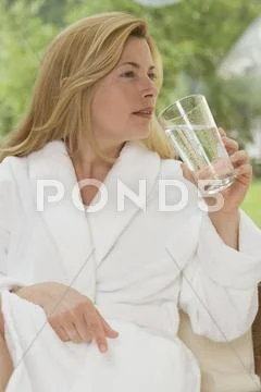 Woman In White Bathrobe Drinking Glass Of Water In Garden