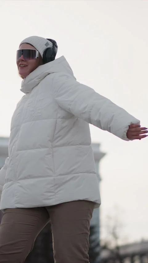 Woman in White Puffer Jacket Dancing Energetically Outdoors with Headphones and Stock Footage 346795932