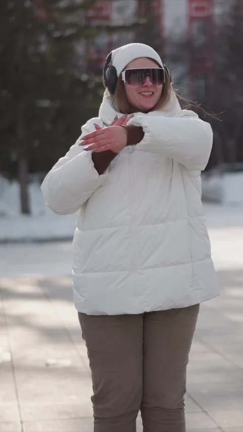 Woman in white puffer jacket gesturing outdoors with headphones and sunglasses Stock Footage 346795609