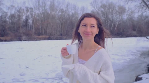 Woman in a white pullover drinks hot drink from a white cup on the bank of the i Vídeos de archivo 169636733