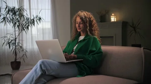 Woman work remotely using laptop sitting at couch.Curly hair girl looking at Stock Footage 212315546