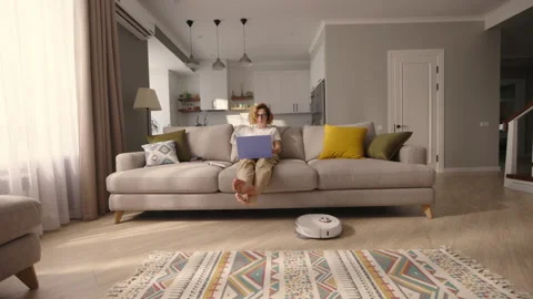Woman working with laptop on sofa while robot vacuum cleaner cleans apartment Stock-Footage 249938870