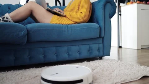 Woman working with laptop on sofa while robot vacuum cleaner cleans Stock Footage 301244304