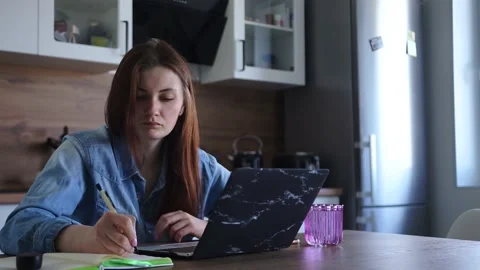 Woman writes notes in notebook, working on laptop at home in kitchen Stock Footage 152876329