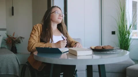 Woman writes notes while sitting in a wheelchair at her home with cookies nearby 스톡 동영상 327558996