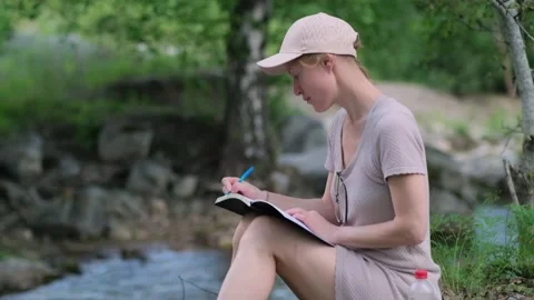 Woman writing in her diary while sitting by water's edge in the forest. Stock Footage 313792087