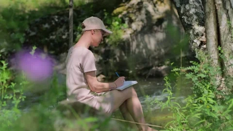 Woman writing in her diary while sitting by water's edge in the forest. Stock Footage 314242885