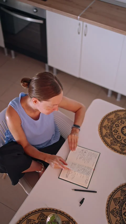 Woman writing in a notebook at a table in a kitchen with patterned coasters Stock Footage 341325783