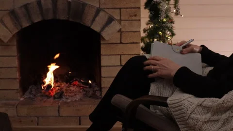 Woman writing in a notebook while sitting in an armchair by a burning fireplace. Stock Footage 326099736