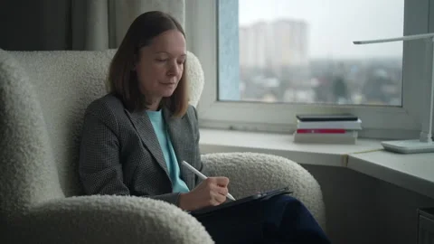 Woman writing notes while sitting by a window with a city view during the day Stock Footage 322164668