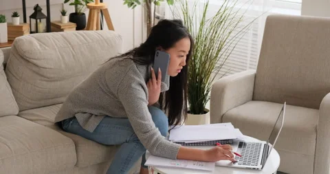 Woman writing notes while using laptop and talking on mobile phone Stock Footage 145295728