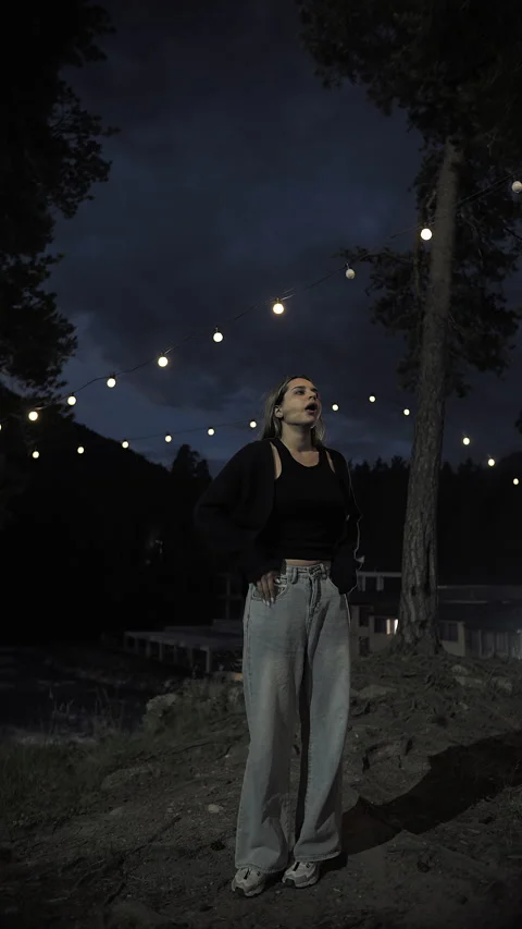 Woman yawning under string lights in a dark forest setting Stock Footage 284335990