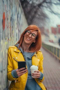 Woman in yellow jacket using smartphone and drinking coffee Stock Photos