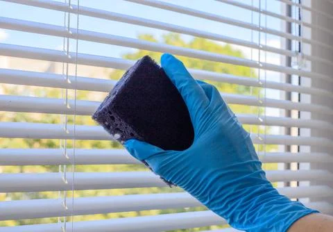 Woman's hand in a blue glove cleans white blinds with a black sponge Stock Photos