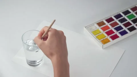 A woman's hand with a drawing brush and watercolor moistens the brush in a glass Stock Footage 202502255