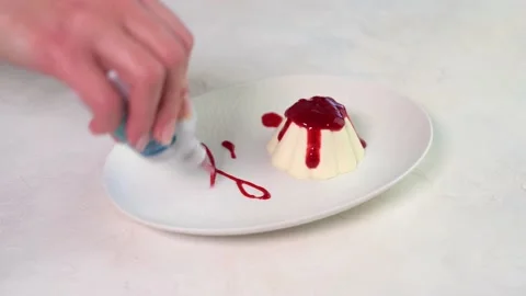 Woman's hand draws a pattern raspberry syrup panna cotta on white plate Stock Footage 149253206