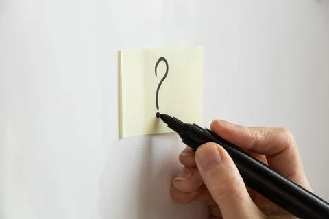 A woman's hand draws a question mark on a yellow sticky note with a black m.. Stock Photos