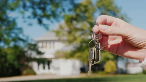 A woman's hand holding house keys with a charming key chain in front of a b.. Stock Footage 334704380