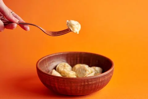 A woman's hand holds a dumpling on a fork in front of a bowl. Orange backgrou Stock Photos