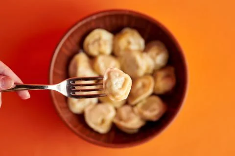 A woman's hand holds a dumpling on a fork in front of a bowl. Orange backgrou Stock Photos