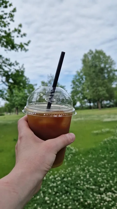 A woman's hand holds a plastic cup of iced coffee against a green park Stock Footage 336782006