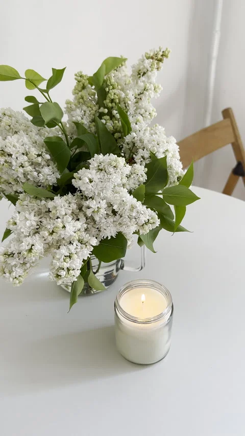 Woman's hand lighting candle in clear glass jar and white lilac bouquet on white Stock Footage 241196201