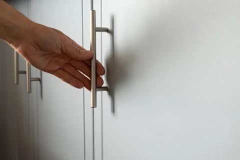 A woman's hand opens a light-colored kitchen cabinet using a metal handle. Th Photos