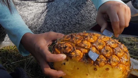 Woman's hand peeling a pineapple on the grass using a kitchen knife. close POV. Video stock 116355724